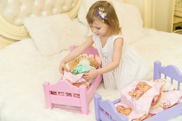 Uma criança pré-escolar ativa, uma menina bonita com um cabelo encaracolado loiro, brinca com suas bonecas, as coloca para dormir em um roubo — Fotografia de Stock