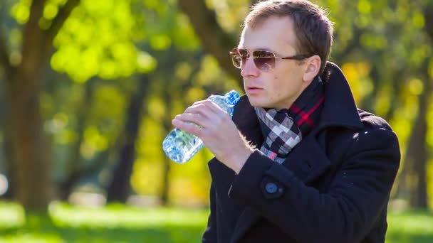 Hombre joven en un abrigo relajante en el parque en el día soleado y agua potable, steadycam disparo — Vídeos de Stock