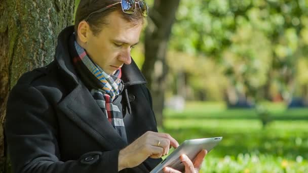 Jonge zakenman met een vacht met tablet pc zit in het park — Stockvideo