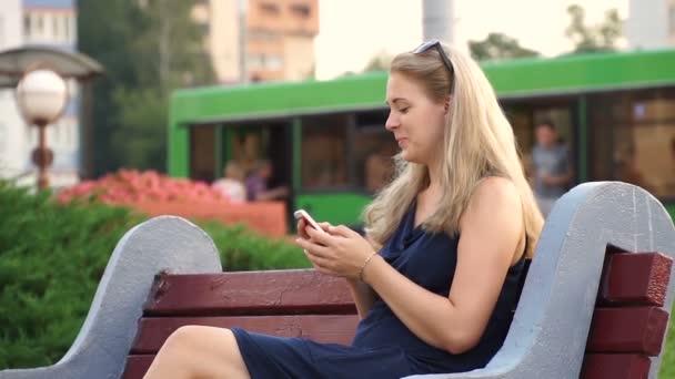 Boldog lány okos telefont használ egy városi parkban, egy padon ülve. — Stock videók