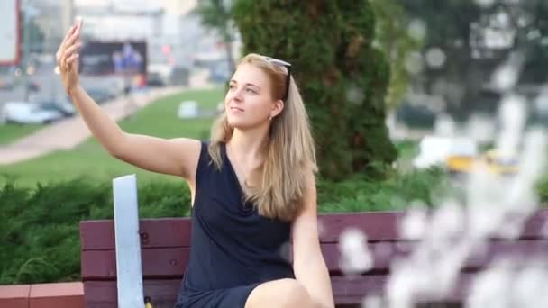 Una chica está tomando fotos de sí misma en el teléfono mientras está sentada en un parque en un banco junto a la fuente — Vídeos de Stock