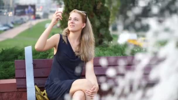 Una ragazza si sta fotografando al telefono mentre è seduta in un parco su una panchina accanto alla fontana — Video Stock