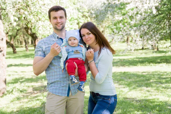 Familia feliz joven caminando al aire libre. Los padres sostienen al niño en las manos y se regocijan. Son felices juntos. Sonrían el uno al otro — Foto de Stock