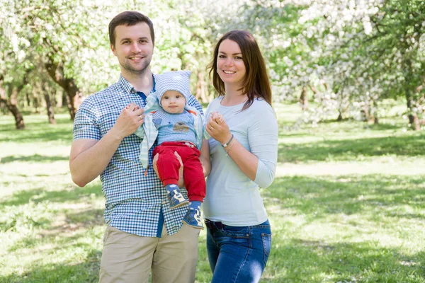 Familia feliz joven caminando al aire libre. Los padres sostienen al niño en las manos y se regocijan. Son felices juntos. Sonrían el uno al otro — Foto de Stock