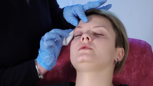Der Spezialist für Permanent Make-up wischt vor dem Eingriff das Gesicht der Patientin mit einem Wattestäbchen ab. Desinfektion. — Stockvideo