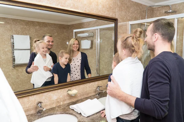 O marido, a esposa e os filhos ficam em frente ao espelho no banheiro do quarto do hotel e sorriem. Família jovem é muito feliz — Fotografia de Stock