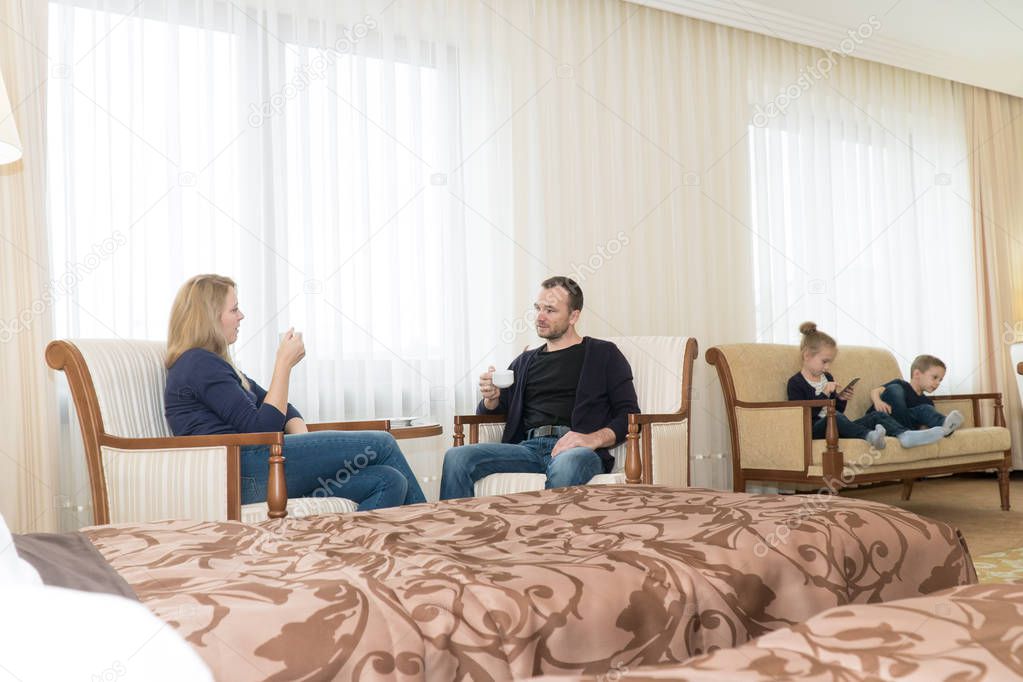 The husband and wife and the children in the hotel room. A children are sitting on the bed, parents on chairs. Young family is very happy