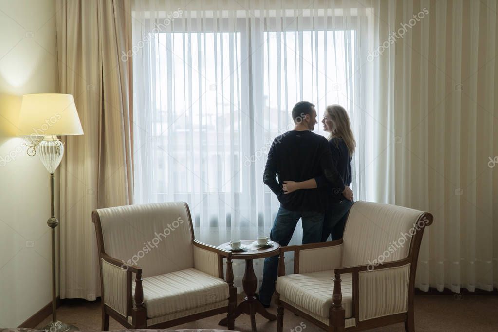 The newlyweds stand at the window of the hotel room and look at each other. Young family is very happy