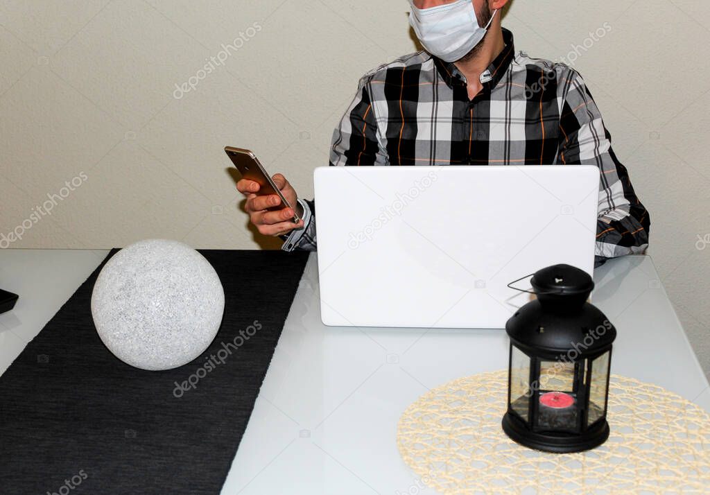 New normality. A man in a protective mask quarantined, working from his living room with his laptop and white smartphone.