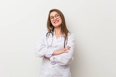 Genç doktor kadın beyaz bir duvara yaslanmış kollarını kavuşturarak gülümsüyor..