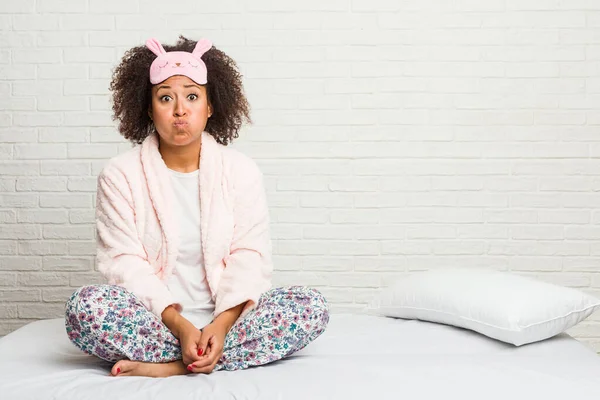 Young african american woman in the bed wearing pijama blows cheeks, has tired expression. Facial expression concept.