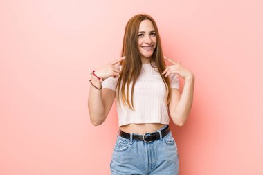 Kızıl saçlı genç kadın pembe bir duvara yaslanmış gülümsüyor, parmaklarını ağzına doğrultuyor..