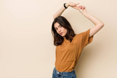 Genç esmer kadın bej bir arka planda kollarını uzatıyor, rahat bir pozisyonda..