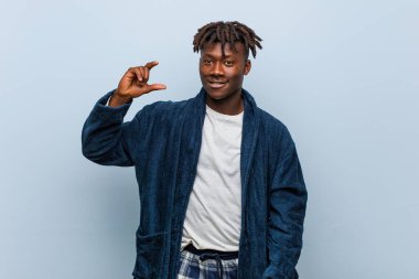 Genç Afrikalı siyah adam pijama giyiyor. Elinde işaret parmaklarıyla küçük bir şey tutuyor, gülümsüyor ve kendinden emin..
