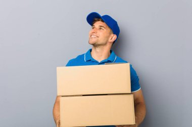 Paketleri teslim eden genç adam kollarını kavuşturup gülümsüyor..