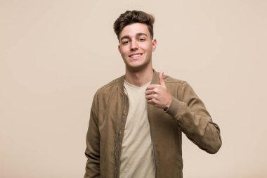 Kahverengi ceketli genç beyaz adam gülümsüyor ve başparmağını kaldırıyor.