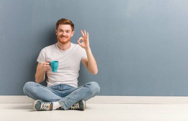 Genç kızıl saçlı öğrenci yerde neşeli ve kendinden emin bir şekilde oturuyor ve iyi bir jest yapıyor. Elinde bir kahve fincanı var..