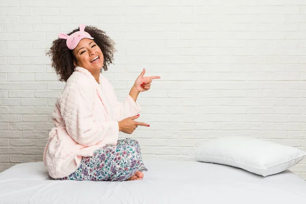 Young african american woman in the bed wearing pijama excited pointing with forefingers away.
