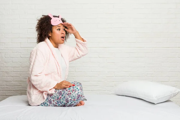 Young african american woman in the bed wearing pijama looking far away keeping hand on forehead.