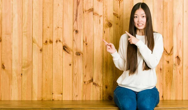 Młoda Chinka Siedząca Drewnianym Miejscu Zszokowana Wskazując Palcem Wskazującym Miejsce — Zdjęcie stockowe