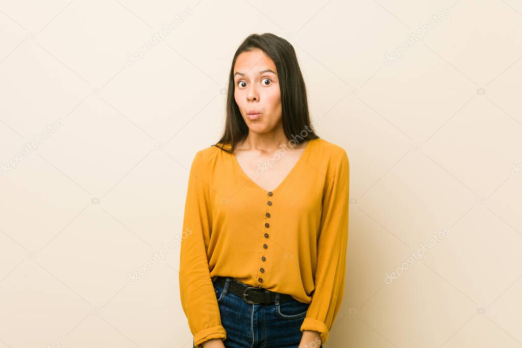 Young hispanic woman against a beige background shrugs shoulders and open eyes confused.