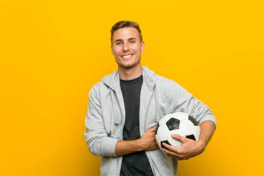Genç, beyaz bir adam elinde futbol topu tutarken kollarını kavuşturup gülümsüyor..