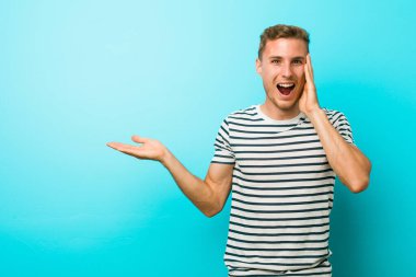 Mavi duvara yaslanmış genç beyaz bir adam, avuç içinde fotokopi alanı tutuyor, el üstünde tutuyor. Şaşırmış ve mutlu..