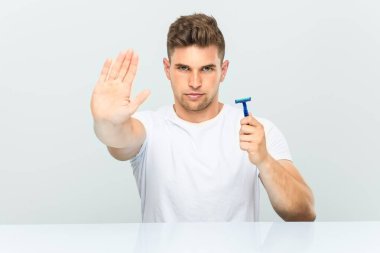Young man holding a razor blade standing with outstretched hand showing stop sign, preventing you.