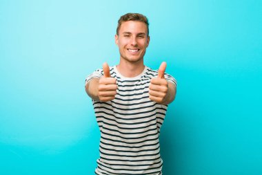 Mavi duvara yaslanmış genç, beyaz bir adam. Başparmakları havada, bir konuda tezahürat yapıyor. Destek ve saygı kavramı..