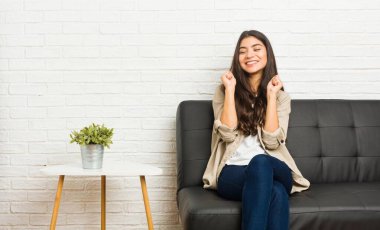 Genç Arap kadın koltukta oturuyor, yumruğunu kaldırıyor, mutlu ve başarılı hissediyor. Zafer konsepti.