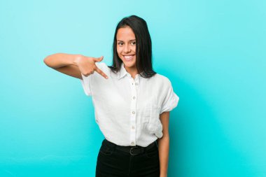 Genç, İspanyol asıllı, havalı bir kadın, mavi bir duvara karşı. Elle bir tişört fotokopisini gösteren, gururlu ve kendinden emin.