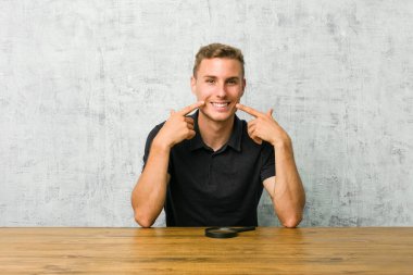 Elinde büyüteç tutan genç yakışıklı adam gülümsüyor, parmaklarını ağzına doğrultuyor..