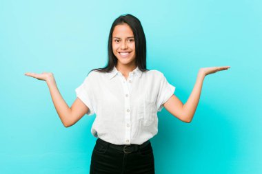 Mavi bir duvara yaslanmış genç Latin asıllı havalı bir kadın kollarıyla ölçeği yapıyor, mutlu ve kendinden emin hissediyor..