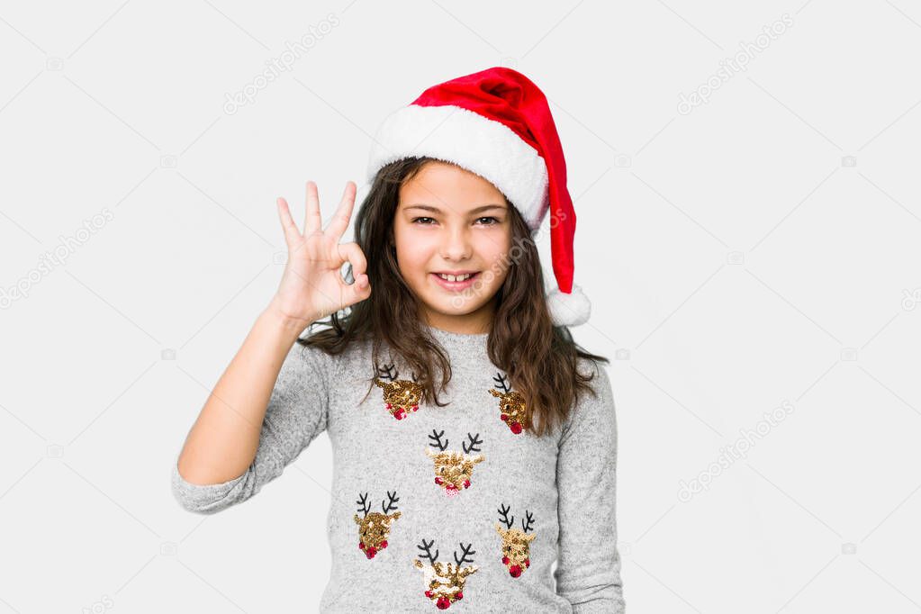 Little girl celebrating christmas day winks an eye and holds an okay gesture with hand.