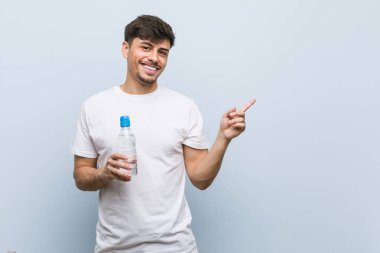 Young hispanic man holding a water bottle smiling cheerfully pointing with forefinger away. clipart