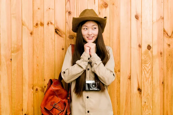 Young chinese traveler woman keeps hands under chin, is looking happily aside.