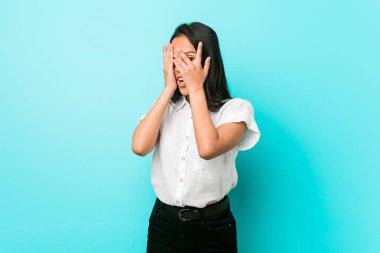 Genç Latin asıllı havalı bir kadın mavi bir duvara yaslanmış korkmuş ve gergin parmaklarıyla göz kırpıyor..