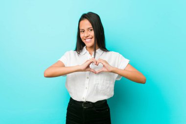 Mavi bir duvara yaslanmış, gülümseyen ve elleriyle kalp şeklini gösteren genç İspanyol bir kadın..