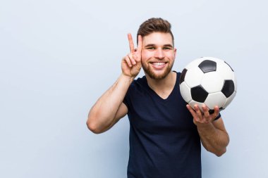 Genç beyaz adam zafer işareti gösteren bir futbol topu tutan ve geniş gülümseyen.