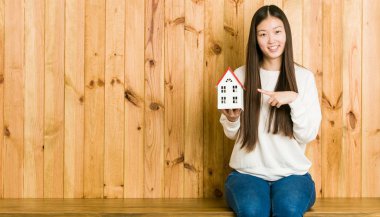 Young asian woman holding a house icon smiling cheerfully pointing with forefinger away.