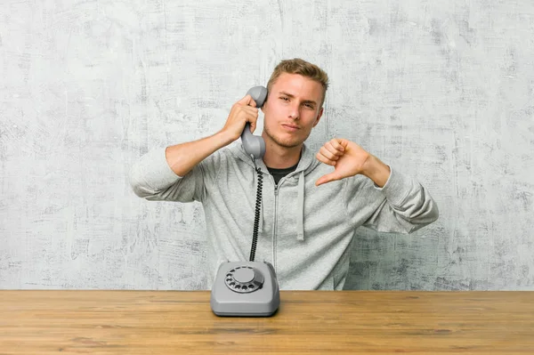 Young man talking on a vintage phone showing thumb down and expressing dislike.