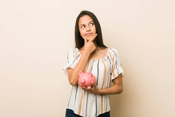 Young Hispanic Woman Holding Piggy Bank Looking Sideways Doubtful Skeptical — Stock Photo, Image