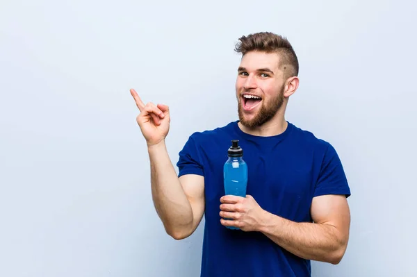 Joven Deportista Caucásico Sosteniendo Una Bebida Isotónica Sonriendo Alegremente Señalando —  Fotos de Stock