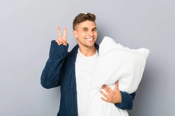勝利の印を示す枕を持っているPijamaを身に着けている若い男と笑顔広く — ストック写真