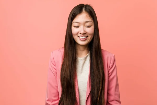 Young business chinese woman wearing pink suit laughs and closes eyes, feels relaxed and happy.