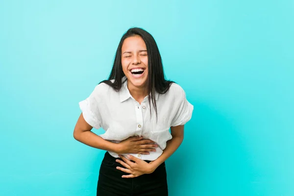 Young hispanic cool woman against a blue wall laughs happily and has fun keeping hands on stomach.