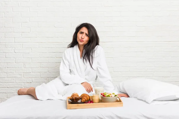 Young curvy woman taking a breakfast on the bed blows cheeks, has tired expression. Facial expression concept.