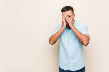 Genç Güney Asyalı adam parmaklarının arasından korkmuş ve gergin bir şekilde göz kırpıyor..