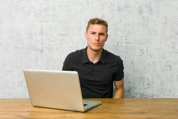 Young entrepreneur working with his laptop on a desk blows cheeks, has tired expression. Facial expression concept.