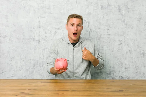 Young man saving money with a piggy bank surprised pointing with finger, smiling broadly.